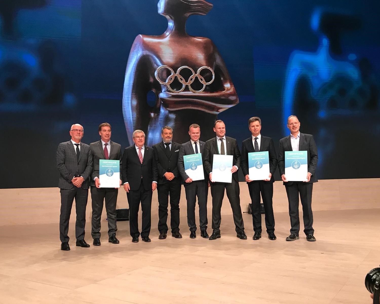 The cities' representatives stand on a stage with their certificate in hand.